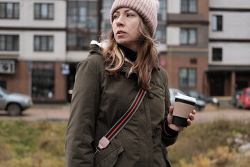 Fototapeta premium Portrait of mature woman in wool hat standing on the urban street, drinking takeaway coffee from paper cup. Middle-aged 40s female walking around the city at cold autumn day. Living in modern district