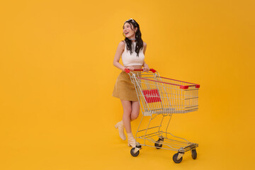 Full length happy excited Asian woman pushing empty shopping cart or shopping trolley grocery walking promotion supermarket isolated on yellow background. © NaMong Productions