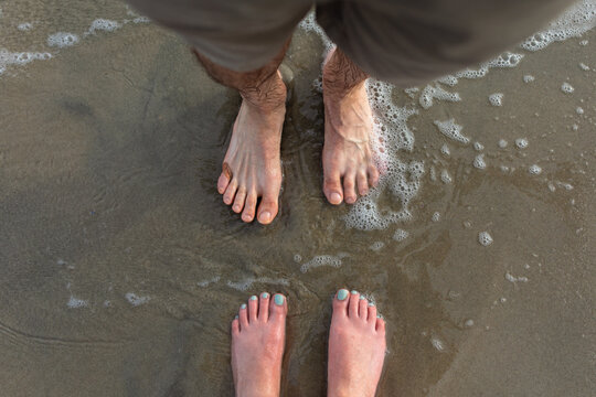 close-up of a white woman's feet and a white man's feet barefooted and facing each other in the sand with ocean waves running over them