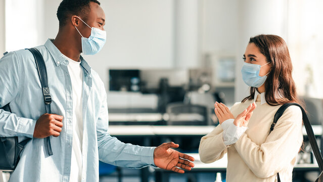 No Handshake, Avoid Contact. Multicultural students wearing protective face masks and backpacks greeting each other. Woman rejects touching, showing stop sign gesture, standing at classroom