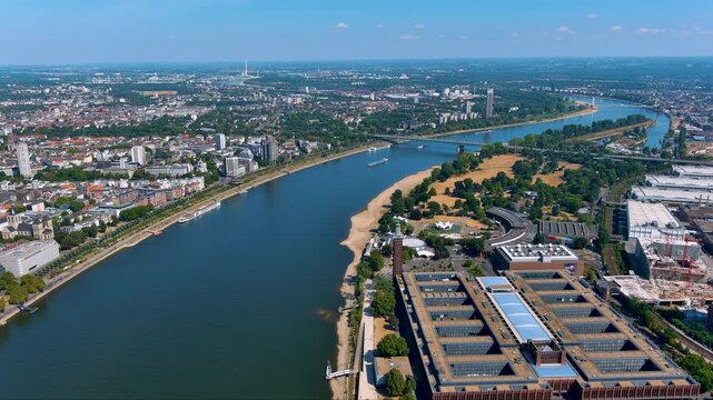 4K drone view of the Rhine curving past Cologne Deutz - Koelnmesse halls, Rheinpark beaches and bridges, cruise boats and skyline framed by green banks on a bright summer day in Germany.