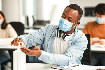 Preventive Measures Concept. Black guy using antibacterial hand sanitizer, pouring it on palms from the bottle, wearing disposable surgical face mask, sitting at desk in classroom during lecture