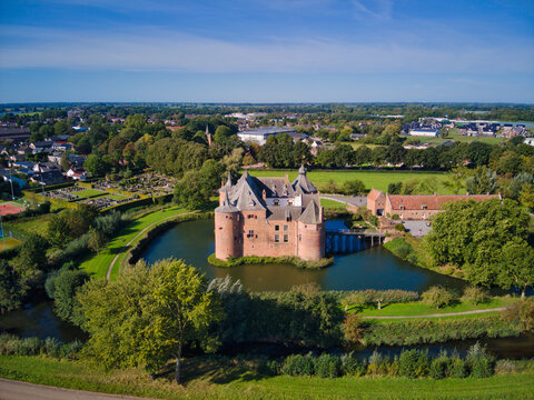 Aerial view of a medieval castle surrounded by a moat, its red brick contrasting with the lush green landscape under a clear blue sky, Helmond, Netherlands.