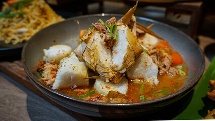 A close-up of traditional Indonesian ketupat (rice cake wrapped in palm leaf) served in a spicy soup with shredded beef, vegetables, and aromatic herbs. A classic Southeast Asian festive dish commonly