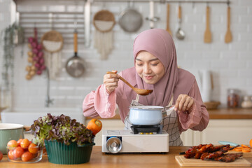 Muslim woman dressed in hijab happy cooking in modern kitchen, focusing on dish in pot with wooden spoon, have fresh vegetables and fruits placed nearby. joyful to scoop up and taste food.