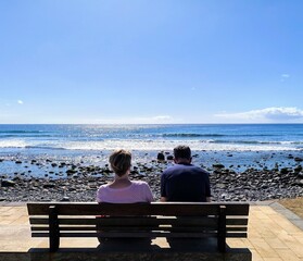 Mature tourist couple sitting and gazing at the sea. Retired people, seen from behind, sitting in front of the sea. Couple sitting and gazing at the horizon, with the sea in the background, on a sunny