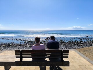 Mature tourist couple sitting and gazing at the sea. Retired people, seen from behind, sitting in front of the sea. Couple sitting and gazing at the horizon, with the sea in the background, on a sunny