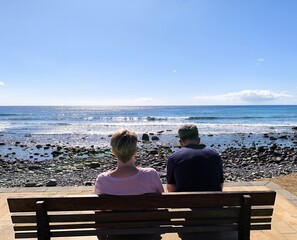 Mature tourist couple sitting and gazing at the sea. Retired people, seen from behind, sitting in front of the sea. Couple sitting and gazing at the horizon, with the sea in the background, on a sunny