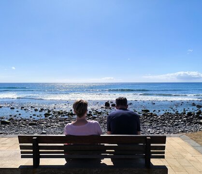 Mature tourist couple sitting and gazing at the sea. Retired people, seen from behind, sitting in front of the sea. Couple sitting and gazing at the horizon, with the sea in the background, on a sunny