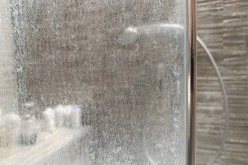 Close-up of a dirty shower glass door with white mineral residue from hard water and soap scum. Blurred shower cabin in the background. Common bathroom cleaning issue.