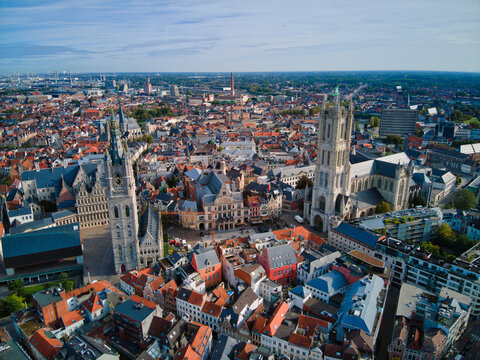 Aerial view of the towering spires of St. Nicholas' Church and St. Bavo's Cathedral rise amidst a sea of red-tiled roofs, creating a striking contrast against the vast blue sky, Ghentent, Flanders.