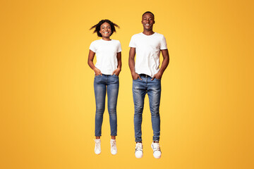 Happy black lovers young man and woman jumping up with hands in pockets, full length shot, orange studio background. Active and lifeful african american couple bouncing together