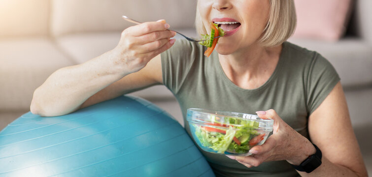 Sports and nutrition concept. Smiling senior lady leaning on fitness ball, eating fresh vegetable salad at home. Athletic mature woman enjoying her diet and exercising program