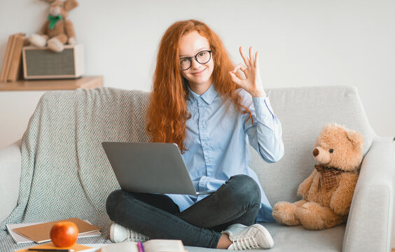 Everything Is Okay. Positive smiling red-haired teenager girl in glasses showing ok sign gesture sitting on the sofa with crossed legs and using laptop, doing homework, recommending online school