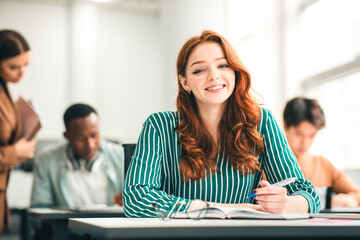 Modern Education Concept. Portrait of smiling ginger red haired female student sitting at desk in classroom at university, taking test or writing notes in her notebook, looking posing at camera