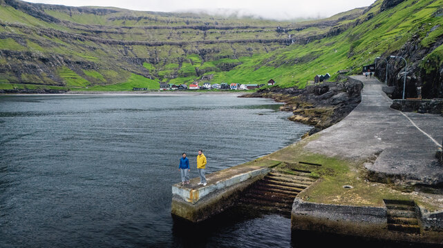Two adventurers stand on a rocky pier overlooking the calm, blue waters of the Faroe Islands. Lush green cliffs tower above, framed by Tjornuvik village