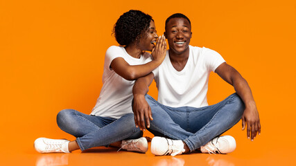 Black woman whispering secrets to her excited boyfriend over orange studio background, empty space. African american woman sharing rumors with her curious man, sitting on floor together, having fun