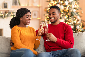 Happy New Year and Christmas celebration. Millennial cheerful african american guy and woman clink glasses of champagne, sit on sofa in living room interior with Xmas tree and glowing garlands