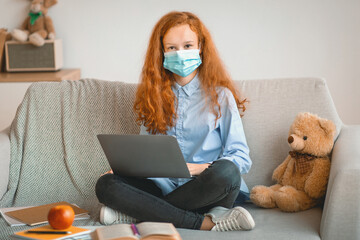 Remote Education During Quarantine, Lockdown. Portrait of girl wearing medical disposable face mask studying at home on laptop computer, sitting on couch and looking at camera, doing homework