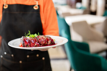 Waiter in orange shirt and black apron serves glazed meat with greens on white plate in stylish restaurant interior with green chairs and tables, shallow depth of field.
