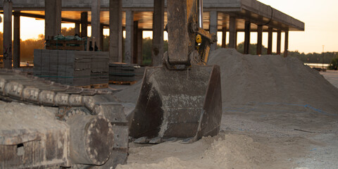 Heavy equipment works diligently at a construction site as the sun sets, with piles of sand and materials visible. The atmosphere reflects industry and progress