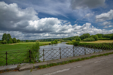Scenic countryside view with river running through green fields, seen from a bridge with metal railing, under dramatic sky filled with large fluffy clouds.