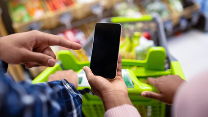 Unrecognizable black couple holding smartphone with empty screen at supermarket, mockup for your mobile app or website. African American guy and lady with cellphone shopping at mall