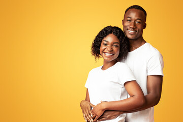 Happy african american young loving couple in white t-shirts bonding and smiling at camera over orange studio background, standing together next to copy space for text or advertisement