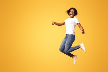 Definitely recommend it. Emotional young black woman jumping in the air and screaming, showing thumbs up, happy african american lady expressing positive emotions on orange studio background