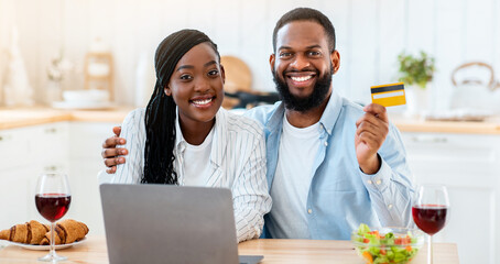 Portrait Of Cheerful Black Lovers With Laptop And Credit Card In Kitchen, Young African American Spouses Making Online Shopping Or Ordering Food Delivery, Sitting At Table And Smiling At Camera