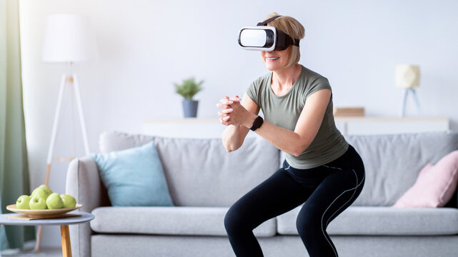 Exercising with virtual reality concept. Athletic mature woman in VR headset doing squats on her home workout. Positive senior lady using modern technologies for domestic training
