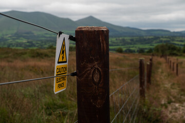 A striking shot of a wooden fence post with a "caution electric fence" sign against a backdrop of rolling hills and a cloudy sky