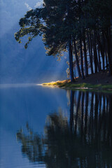 morning sunlight streaming through pine trees with mist over Pang Ung lake in Mae Hong Son, Thailand, reflecting on calm water creating a serene and peaceful atmosphere