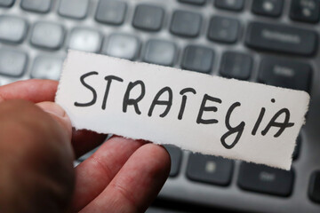 A hand holding a paper note with the word strategia stands out against a blurred keyboard background, symbolizing strategy, planning, and organization in the workplace.