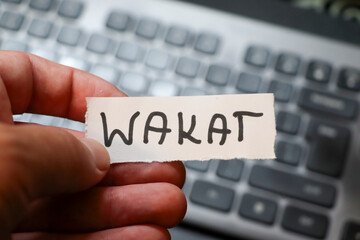 A hand holding a paper strip labeled WAKAT over a blurred computer keyboard. Represents the concept of vacation or work-life balance in modern life.