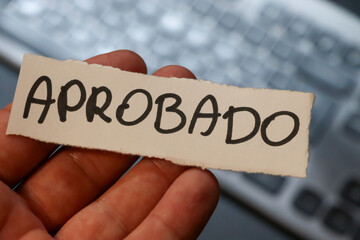 Hand holding a piece of paper with the word APROBADO, symbolizing approval or acceptance, against a blurred keyboard background, ideal for business and educational contexts.