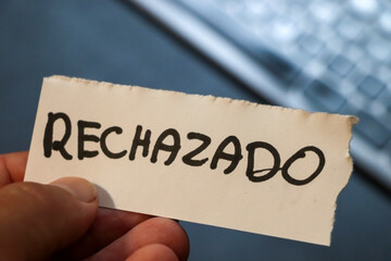 A hand holds a torn paper note with the word RECHAZADO written in bold text, symbolizing rejection or denial, with a blurred keyboard in the background.