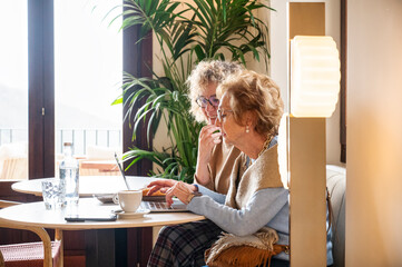 Elderly woman receiving digital assistance from younger companion while learning to use a laptop in cozy café, highlighting senior education and intergenerational support