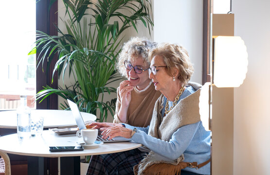 Elderly woman learning to use laptop with help from younger mentor in bright café, focused on digital skills development and intergenerational technology support - Powered by Adobe