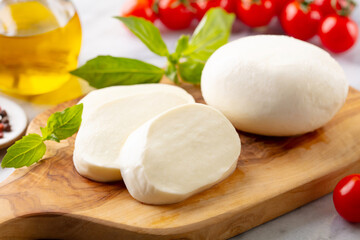Fresh mozzarella cheese with cherry tomatoes and basil leaf on a cutting board. White marble background. Close up.