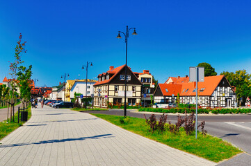 Ustka ,Polska - Piękny krajobraz miejski z wybrukowanym chodnikiem, tradycyjnymi budynkami z pomarańczowymi dachami i bujną zielenią w jasny, słoneczny dzień. © Pol Badger