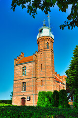 Ustka - Polska -Latarnia morska. Tradycyjna, czerwona, ceglana latarnia morska otoczona drzewami i żywopłotami w słoneczny, bezchmurny dzień. Latarnia ozdobiona jest detalami architektonicznymi. © Pol Badger