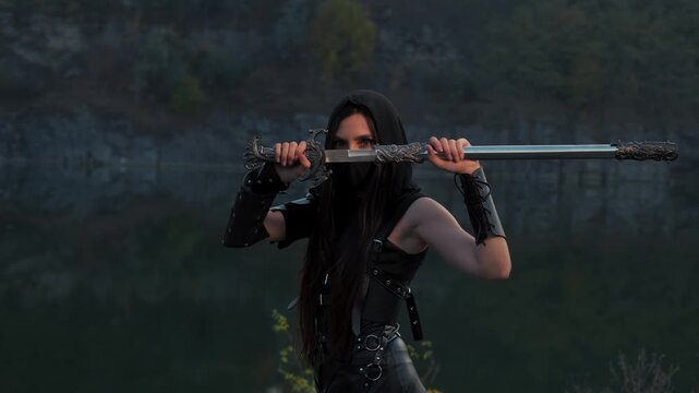 close-up portrait fantasy woman warrior assassin holding dagger in hands, hiding face behind mask. Armed girl, blue eyes. Black leather costume hood on head. Ninja soldier with knife sword. 4k footage