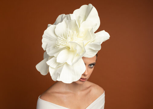 Mysterious female model in white floral headpiece covering part of her face, gazing downward. Conceptual portrait on orange background, elegance and anonymity.