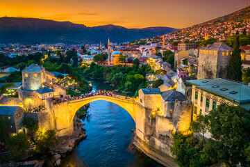 Drone photo of Stari Most in Mostar at sunset showing the historic stone bridge, Neretva river, old town architecture and tourists enjoying the warm glow of golden hour light