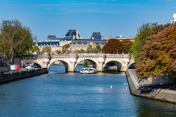 Autumnal cityscape along the Seine, featuring a historic bridge, riverboats, and the iconic Louvre Museum.