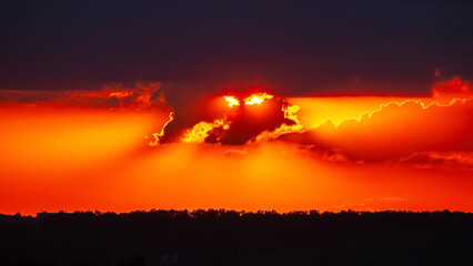Sunset over the horizon with vibrant colors and dramatic clouds near a wooded landscape