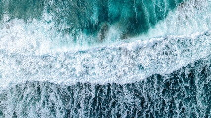 Aerial drone view of ocean surf crashing onto the shore. Turquoise water with white foam creates abstract natural patterns – a dynamic top-down shot of waves and sea textures.