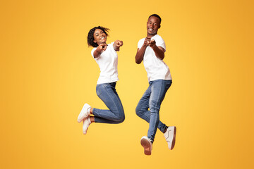 Happy black lovers attractive young man and woman wearing jeans and white t-shirts posing on orange studio background, jumping in the air and pointing fingers at camera. People lifestyle concept