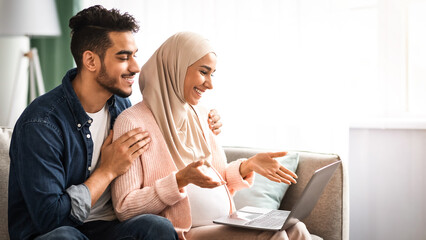 Cheerful Pregnant Islamic Spouses With Laptop Making Video Call At Home, Happy Muslim Couple Having Online Meeting With Friends Or Family Via Web Conference On Computer, Talking At Camera And Smiling